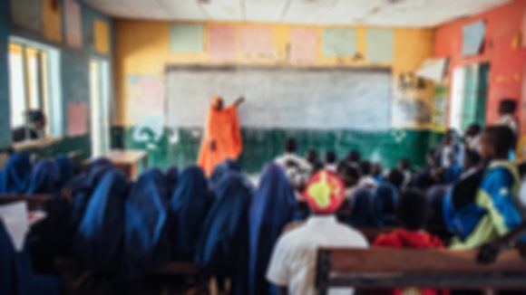 A classroom with a teacher pointing to a whiteboard. A blurred filter has been applied to suggest what the board would look like for someone with a refractive error.