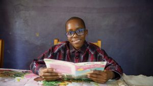 Jayden sits at a table reading. He is wearing his glasses.