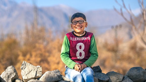 A boy wearing a football jersey sits on a stone wall smiling. He is wearing a pair of glasses.