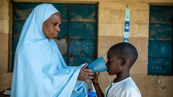 A boy drinks from a cup of water, held by an older woman, after taking medication to treat disease.