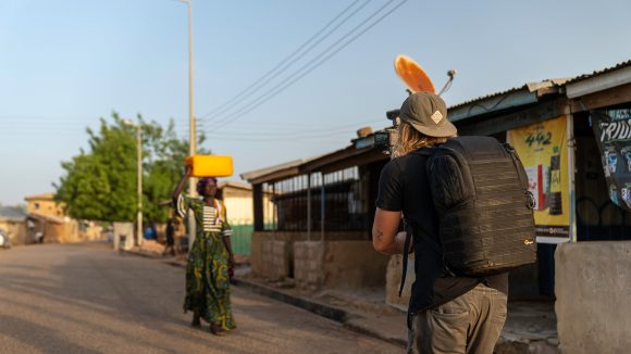 A cameraman walks backwards to capture a woman walking down a road. She is carrying a container on her head.