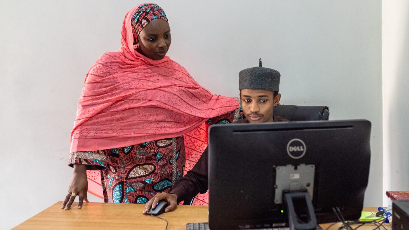 Aisha, a young woman, stands behind Usman, a young man, who is sitting at a computer.