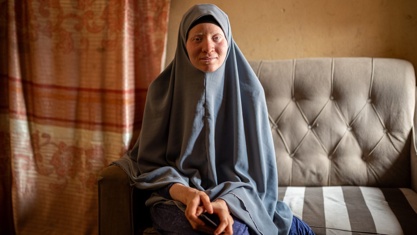 Saudat, who wears a grey hijab, sits on a sofa and smiles warmly.