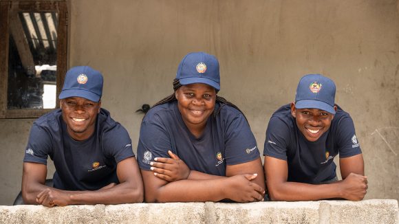 Two men and a woman wearing uniforms from the Mozambique Ministry of Health lean on a wall. They are smiling broadly.