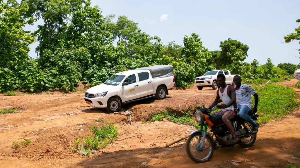 Two men ride on a motorbike down a dusty track. Two 4x4 vehicles drive alongside them.