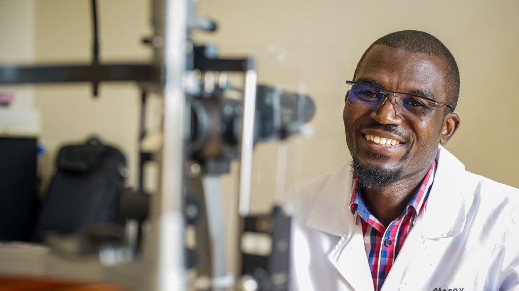 A male health worker smiles while sitting in front of an autorefractor.